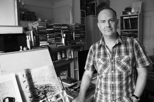 Mark-Buckingham-studio-Turnham-Green-London-W4-standing-by-board-grey-pic#1-30th-July-2015